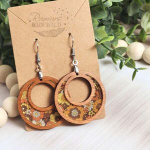 May include: Wooden hoop earrings with a floral pattern in shades of orange, yellow, and brown. The earrings have a layered design with a smaller inner circle. The earrings are displayed on a brown card with the text "Rumors Run Wild".