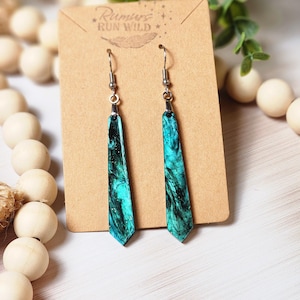 May include: Teal and black marbled dangle earrings with silver hooks. The earrings are on a brown card with the text "Rumours Run Wild" printed on it.