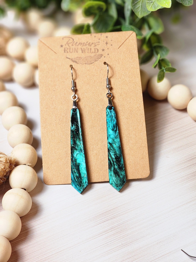 May include: Teal and black marbled dangle earrings with silver hooks. The earrings are on a brown card with the text "Rumours Run Wild" printed on it.