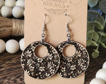 Puede incluir: Un par de pendientes redondos de madera con un patrón floral. Los pendientes son negros y blancos y tienen un gancho plateado.