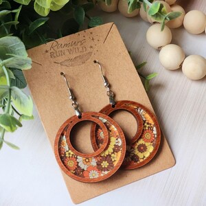 May include: Wooden hoop earrings with a floral pattern in shades of orange, yellow, and brown. The earrings are displayed on a brown card with the text "Rumurs Run Wild". The earrings have silver-colored ear wires.
