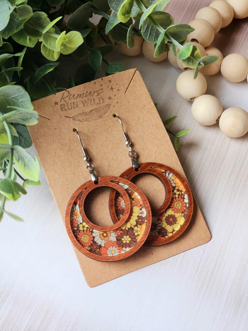 May include: Wooden hoop earrings with a floral pattern in shades of orange, yellow, and brown. The earrings are displayed on a brown card with the text "Rumurs Run Wild". The earrings have silver-colored ear wires.