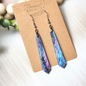 May include: A pair of dangle earrings with a colorful, abstract design. The earrings are long and slender, featuring shades of blue, purple, and black. They hang from silver-colored hooks and are displayed on a brown card with the text "Rumors Run Wild".