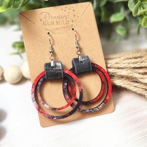 Puede incluir: Pendientes de aro con un dise&ntilde;o en capas. Los pendientes presentan un patr&oacute;n veteado rojo, negro y blanco en los aros, con un cuadrado negro similar al cuero en la parte superior. Los pendientes se exhiben en una tarjeta marr&oacute;n con el texto "Rumors Run Wild".