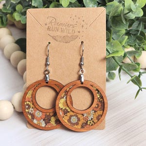 May include: Wooden hoop earrings with a floral pattern in shades of yellow, orange, and brown. The earrings have a circular design with a smaller inner circle. The earrings are displayed on a brown card with the text "Rumors Run Wild".