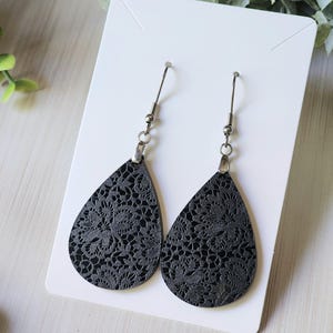 May include: A pair of black teardrop-shaped earrings with a delicate floral pattern. The earrings are made of leather and have silver hooks.