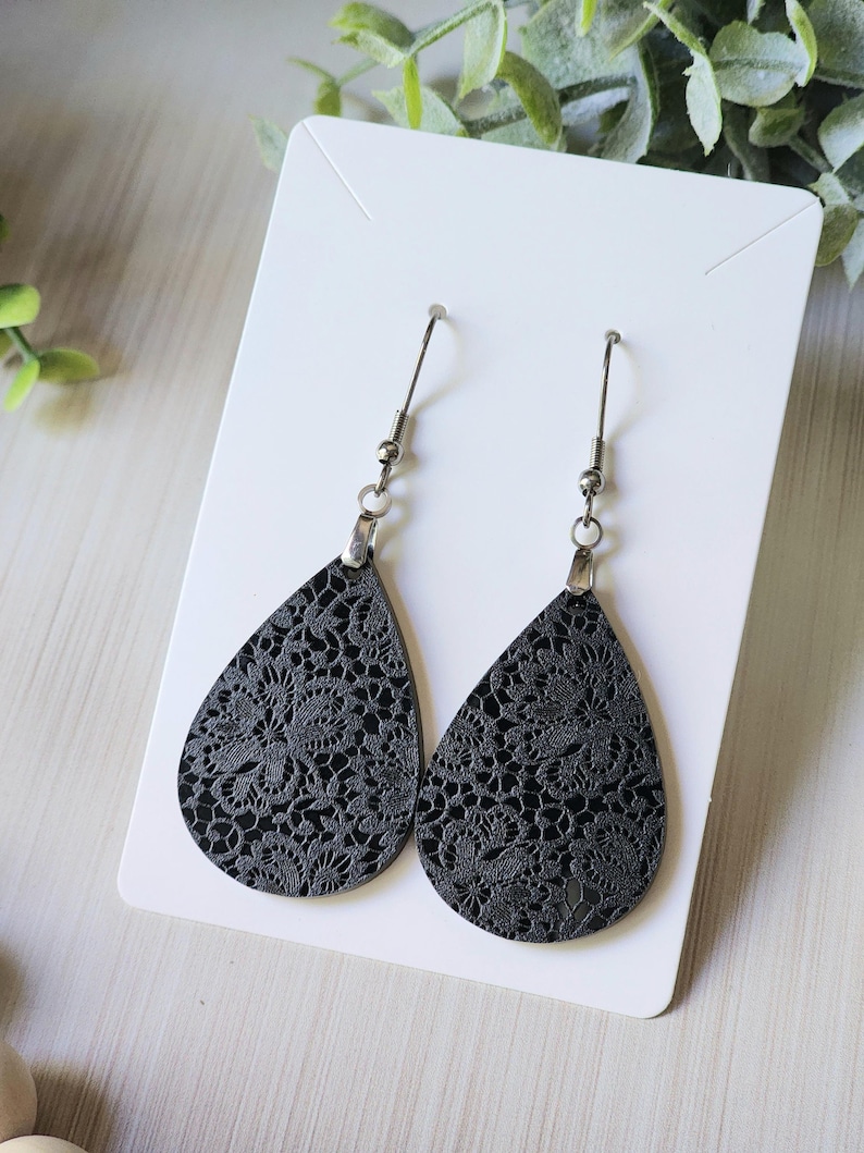 May include: A pair of black teardrop-shaped earrings with a delicate floral pattern. The earrings are made of leather and have silver hooks.