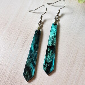 May include: A pair of dangle earrings with a dark green and black marbled design with gold glitter. The earrings have silver hooks.