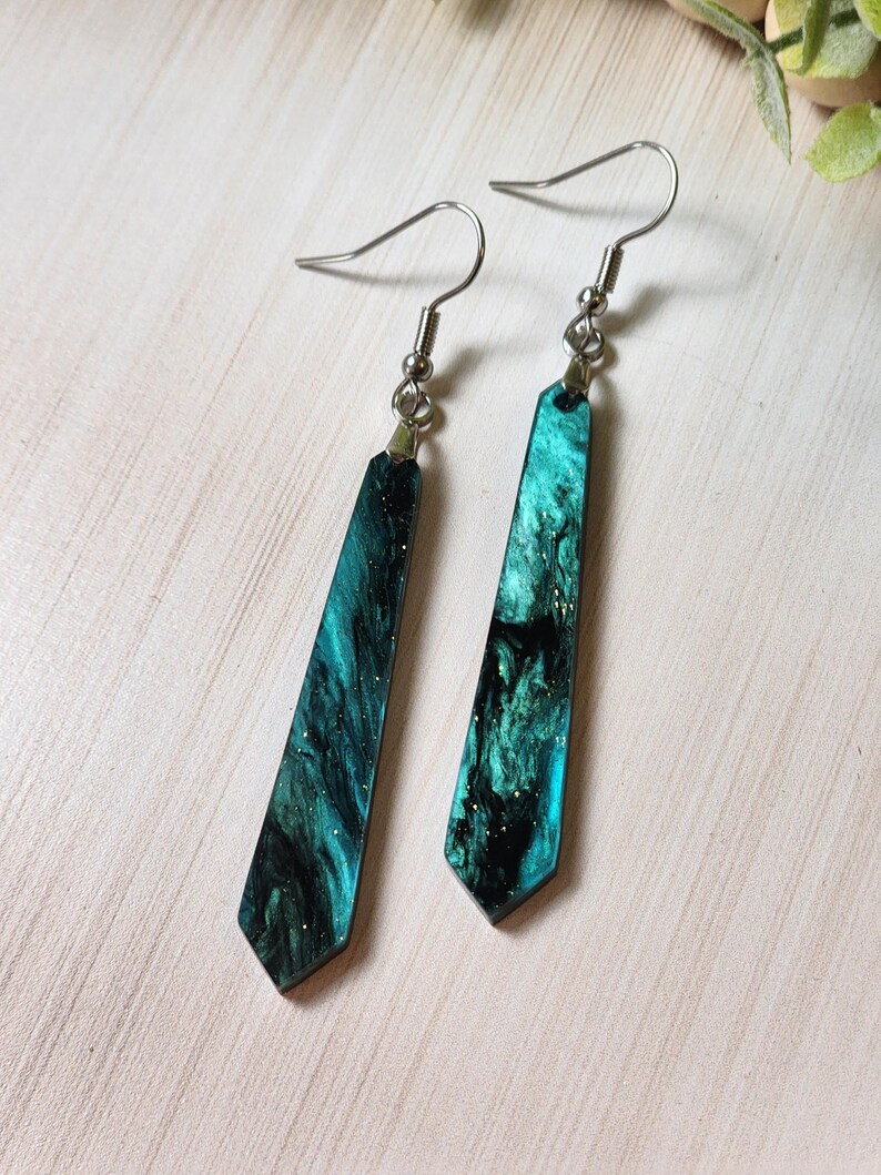 May include: A pair of dangle earrings with a dark green and black marbled design with gold glitter. The earrings have silver hooks.