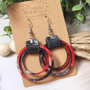 Puede incluir: Pendientes de aro con un dise&ntilde;o veteado rojo, negro y blanco. Los pendientes tienen un detalle de cuero negro y se muestran en una tarjeta marr&oacute;n con el texto "Rumors Run Wild".