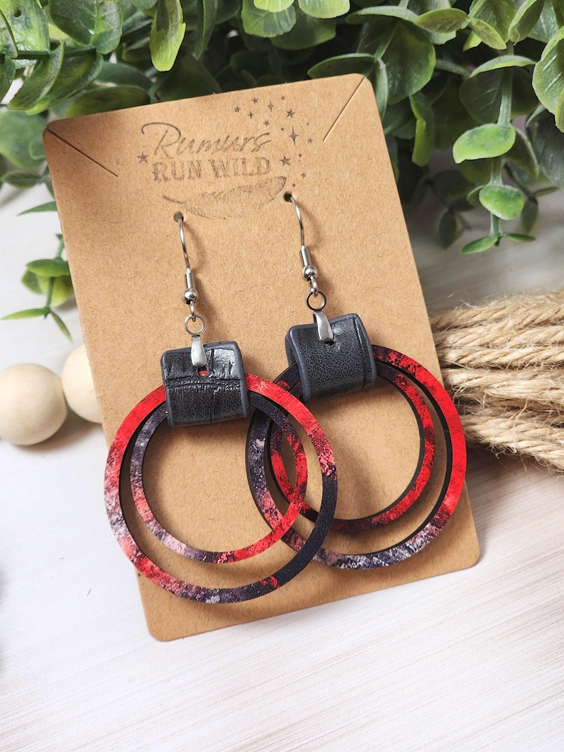 Puede incluir: Pendientes de aro con un dise&ntilde;o veteado rojo, negro y blanco. Los pendientes tienen un detalle de cuero negro y se muestran en una tarjeta marr&oacute;n con el texto "Rumors Run Wild".