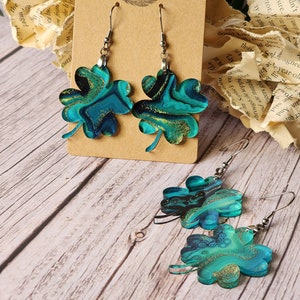 May include: A pair of dangle earrings featuring a four-leaf clover design. The clovers are crafted from green and blue resin with gold glitter accents. The earrings are suspended from silver hooks.