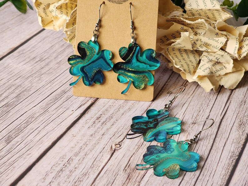 May include: A pair of dangle earrings featuring a four-leaf clover design. The clovers are crafted from green and blue resin with gold glitter accents. The earrings are suspended from silver hooks.
