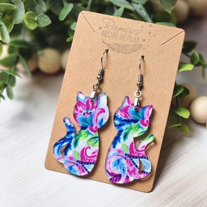 Peut inclure: Boucles d'oreilles en forme de chat avec un motif floral coloré rose, bleu et vert. Les boucles d'oreilles sont présentées sur une carte marron avec le texte "Rumors Run Wild". Elles ont des crochets argentés.