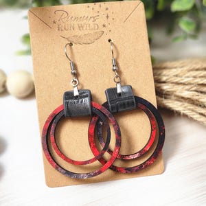 Puede incluir: Pendientes de aro con un dise&ntilde;o veteado rojo y negro. Los pendientes tienen una parte superior rectangular negra y ganchos plateados. Se muestran en una tarjeta marr&oacute;n con el texto "Rumors Run Wild".