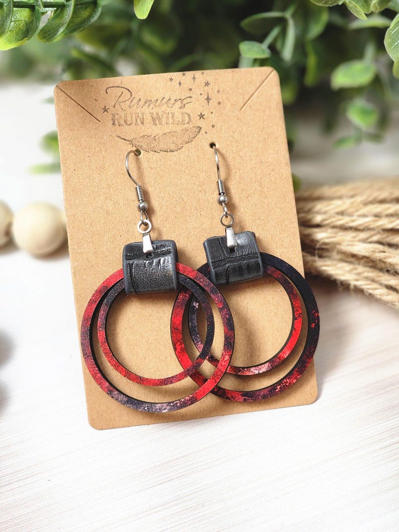 Puede incluir: Pendientes de aro con un dise&ntilde;o veteado rojo y negro. Los pendientes tienen una parte superior rectangular negra y ganchos plateados. Se muestran en una tarjeta marr&oacute;n con el texto "Rumors Run Wild".