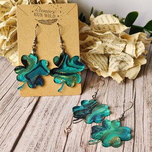 May include: A pair of dangle earrings featuring a shamrock design. The shamrocks are made from green and gold resin with a marbled effect. The earrings are displayed on a brown card with the text "Rumurs RUN WILD".