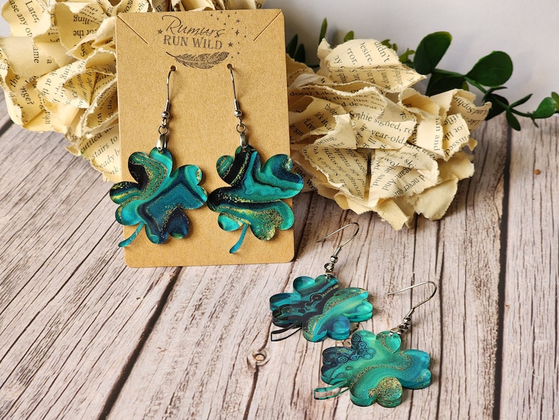 May include: A pair of dangle earrings featuring a shamrock design. The shamrocks are made from green and gold resin with a marbled effect. The earrings are displayed on a brown card with the text "Rumurs RUN WILD".