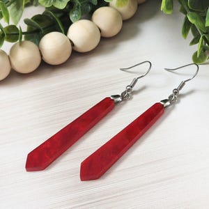 May include: A pair of red, geometric earrings with silver-colored hooks. The earrings have a marbled appearance and a long, slender shape. The background includes greenery and wooden beads, suggesting a handcrafted jewelry theme.