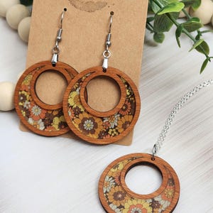 May include: Wooden hoop earrings and a matching pendant necklace. The earrings and pendant feature a floral pattern in shades of orange, yellow, and brown. The earrings hang from silver-colored hooks, and the necklace has a silver chain. The text "Rumors Run Wild" is visible on the backing.