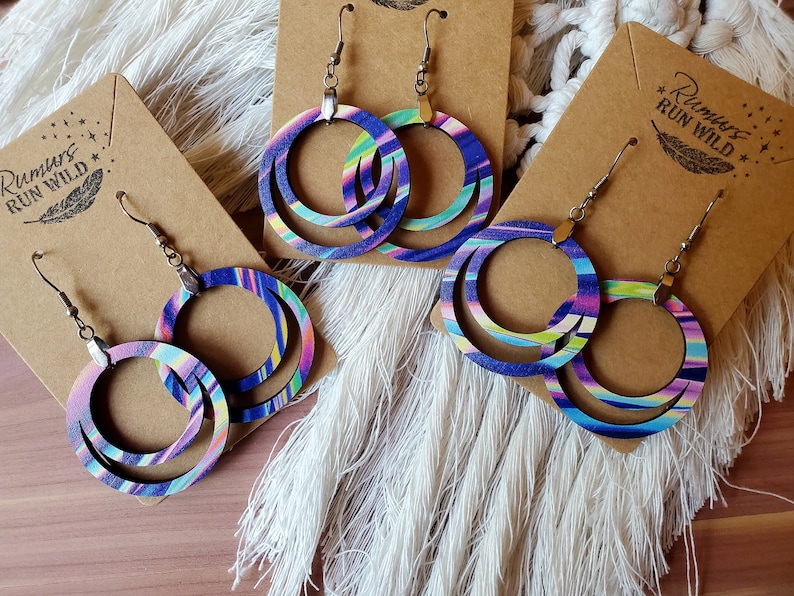 May include: Three pairs of colorful wooden earrings with a layered circle design. The earrings are painted with a blue, purple, pink, and green striped pattern. The earrings are displayed on brown card stock with the text "Rumours Run Wild" printed on the card.