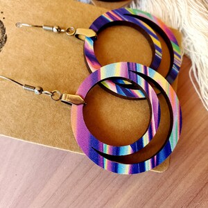 May include: A pair of colorful wooden hoop earrings with a layered design. The earrings are purple, blue, pink, yellow, and green. The earrings have silver hooks.