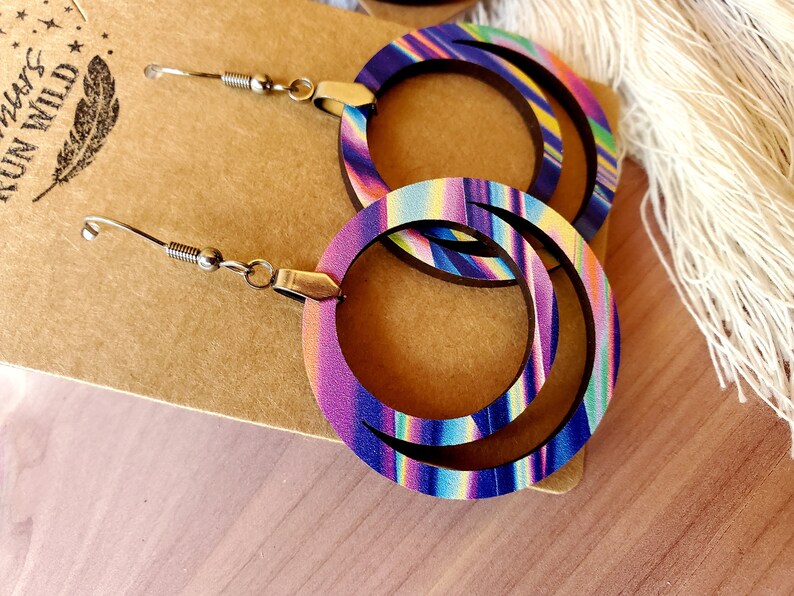 May include: A pair of colorful wooden hoop earrings with a layered design. The earrings are purple, blue, pink, yellow, and green. The earrings have silver hooks.