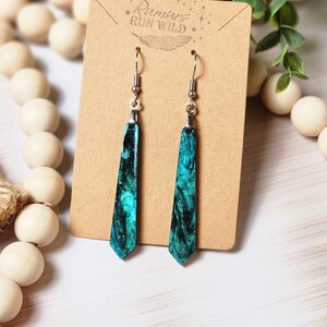 May include: A pair of teal and black dangle earrings with a silver hook. The earrings are on a brown card with the text "Run Wild" in white.