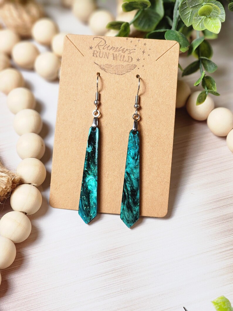 May include: A pair of teal and black dangle earrings with a silver hook. The earrings are on a brown card with the text "Run Wild" in white.