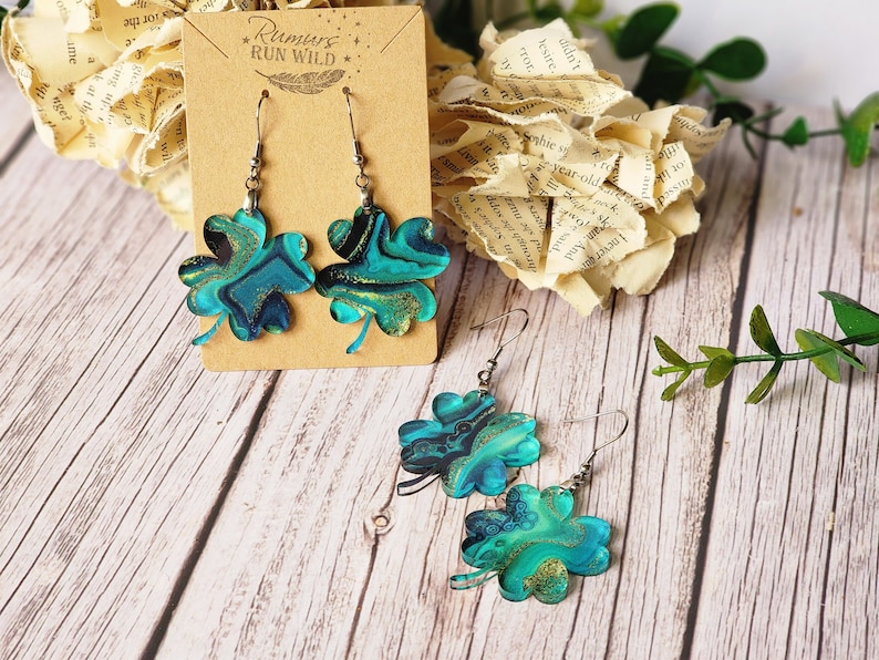 May include: A pair of earrings featuring four-leaf clover charms with a blue, green, and gold marbled design. The earrings are on a brown card with the text "Rumurs RUN WILD".