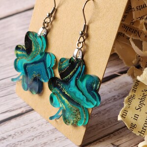 May include: A pair of green and gold clover-shaped earrings with silver hooks. The clovers have a marbled pattern with swirls of green, gold, and black.
