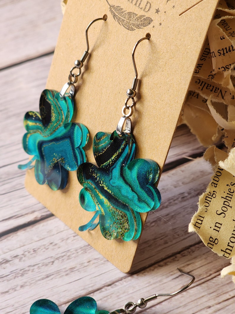 May include: A pair of green and gold clover-shaped earrings with silver hooks. The clovers have a marbled pattern with swirls of green, gold, and black.