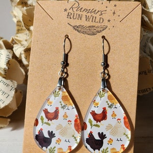 May include: A pair of teardrop-shaped earrings with a clear background and a pattern of colourful chickens. The earrings are silver and have a small silver ball at the top. The card behind the earrings says "Rumors Run Wild" with a feather design.