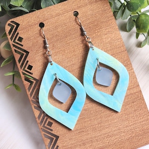 May include: A pair of dangle earrings with a blue and green marbled design. Each earring has a large, open, diamond-shaped frame with a smaller square shape suspended inside. The earrings hang from silver-colored hooks.