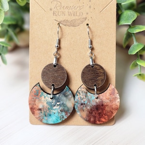 May include: Wooden earrings with a dark brown top circle and a crescent-shaped bottom piece. The bottom piece has a turquoise, copper, and gold-colored design. The earrings hang from silver-colored hooks. The text "Rumors Run Wild" is printed on the backing card.