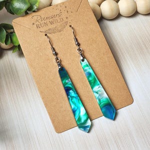 May include: A pair of dangle earrings with a marbled design in shades of blue, green, and white. The earrings are attached to silver-colored fish hook ear wires and are displayed on a brown card with the text "Rumurs Run Wild".
