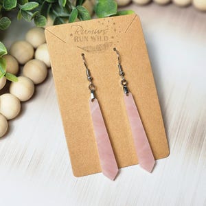 May include: A pair of pink, geometric earrings with silver-colored hooks, displayed on a brown card. The earrings have a long, slender shape with a pointed bottom. The card has the text "Rumure Run Wild".