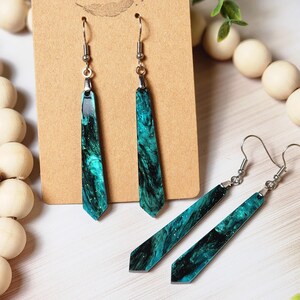 May include: A pair of long, teal and black resin earrings with silver hooks. The earrings are shaped like a long, thin rectangle with a pointed end. The resin has a marbled effect with black and teal swirls. The earrings are on a brown card with the text "Rumours Run Wild" printed on it.