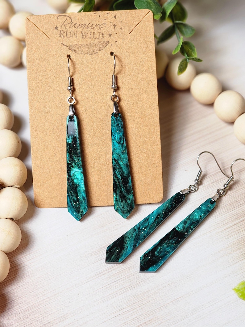 May include: A pair of long, teal and black resin earrings with silver hooks. The earrings are shaped like a long, thin rectangle with a pointed end. The resin has a marbled effect with black and teal swirls. The earrings are on a brown card with the text "Rumours Run Wild" printed on it.