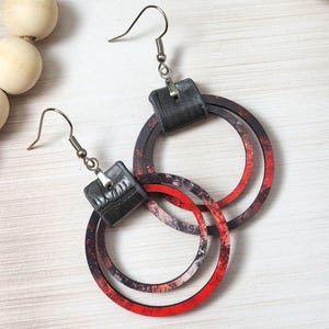 Puede incluir: Un par de pendientes colgantes con una parte superior cuadrada negra y tres aros circulares superpuestos. Los aros tienen un dise&ntilde;o veteado rojo y negro. Los pendientes est&aacute;n suspendidos de ganchos para la oreja de color plateado.