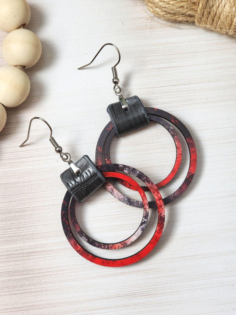 Puede incluir: Un par de pendientes colgantes con una parte superior cuadrada negra y tres aros circulares superpuestos. Los aros tienen un dise&ntilde;o veteado rojo y negro. Los pendientes est&aacute;n suspendidos de ganchos para la oreja de color plateado.