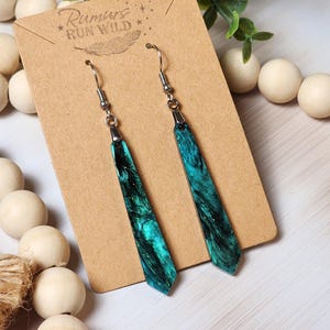 May include: Teal and black resin dangle earrings with silver hooks. The resin has a marbled effect with glitter. The earrings are on a brown card with the text "Rumours Run Wild" in white.