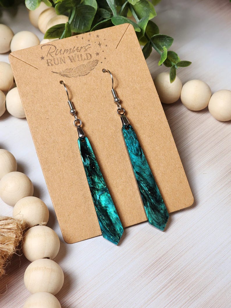 May include: Teal and black resin dangle earrings with silver hooks. The resin has a marbled effect with glitter. The earrings are on a brown card with the text "Rumours Run Wild" in white.