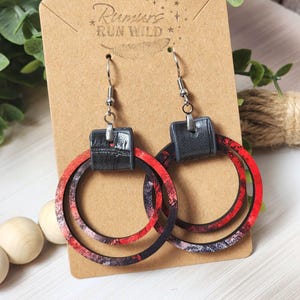 Puede incluir: Pendientes de aro con un dise&ntilde;o veteado rojo, negro y blanco. Los pendientes tienen un cuadrado negro con efecto cuero en la parte superior y ganchos plateados. Se muestran en una tarjeta marr&oacute;n con el texto "Rumors Run Wild".