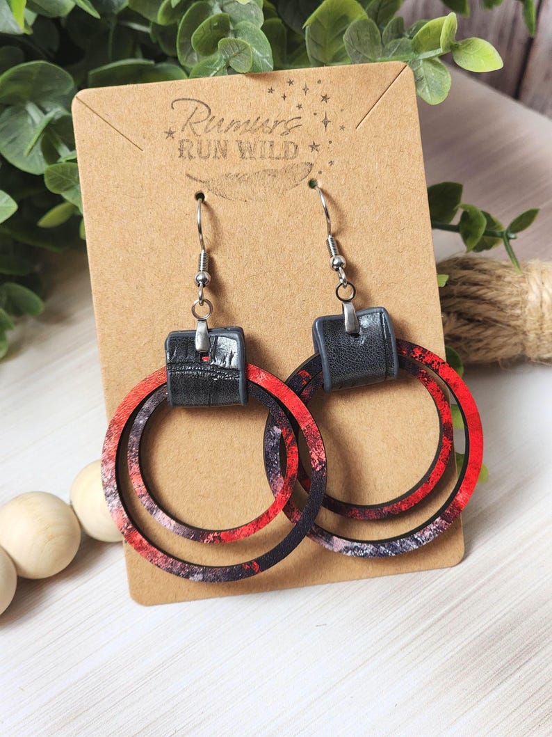 Puede incluir: Pendientes de aro con un dise&ntilde;o veteado rojo, negro y blanco. Los pendientes tienen un cuadrado negro con efecto cuero en la parte superior y ganchos plateados. Se muestran en una tarjeta marr&oacute;n con el texto "Rumors Run Wild".