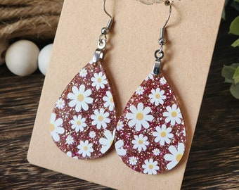 May include: A pair of dangle earrings with a pink glitter background and white daisy print. The earrings are on a brown card with the text "Run Wild" in white.