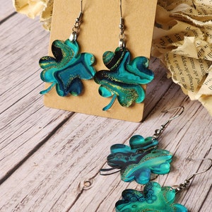 May include: A pair of earrings featuring a four-leaf clover design. The clovers are made of a blue and green marbled material with gold glitter accents. The earrings are hanging from silver hooks.