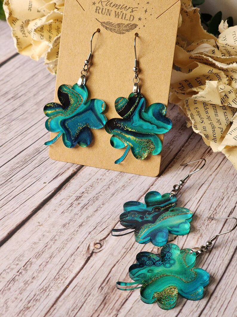 May include: A pair of earrings featuring a four-leaf clover design. The clovers are made of a blue and green marbled material with gold glitter accents. The earrings are hanging from silver hooks.
