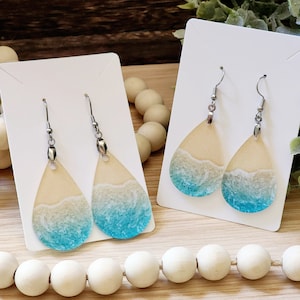 May include: A pair of teardrop-shaped earrings with a beach scene design. The earrings are made of resin and feature a light blue ocean with white foam and a tan beach.