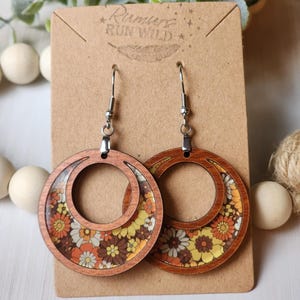 May include: Wooden hoop earrings with a retro floral design in shades of yellow, orange, brown, and white. The earrings are attached to a brown card with the text "Rumurs Run Wild".